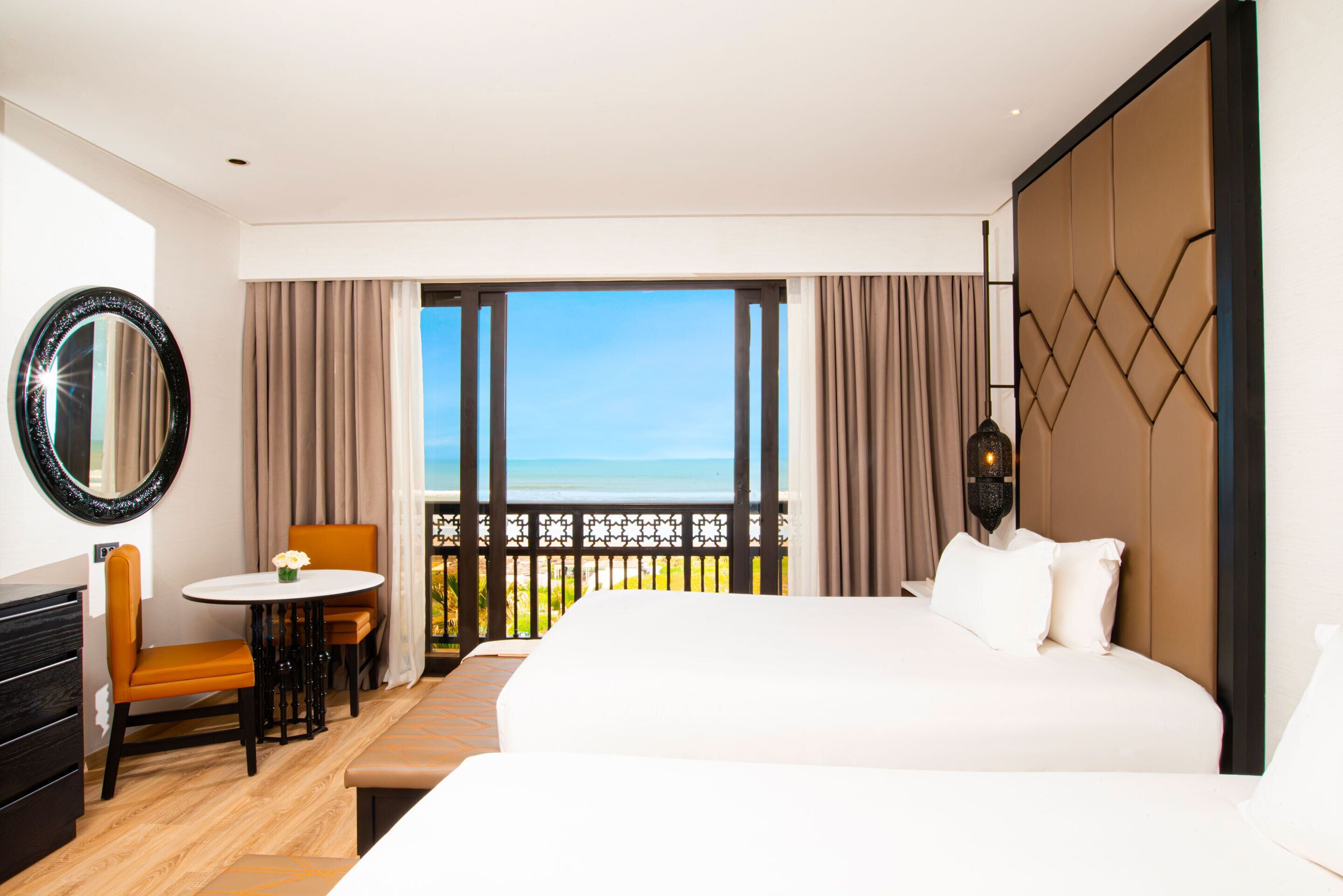 A hotel room with a bed, a desk with a chair, a round mirror, and a view of the ocean.