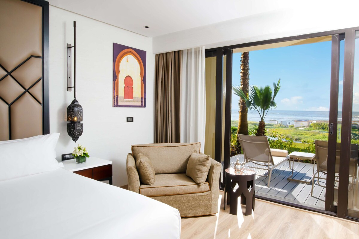 A hotel room with a bed, a sofa, a small table, and a balcony with a view of the ocean.