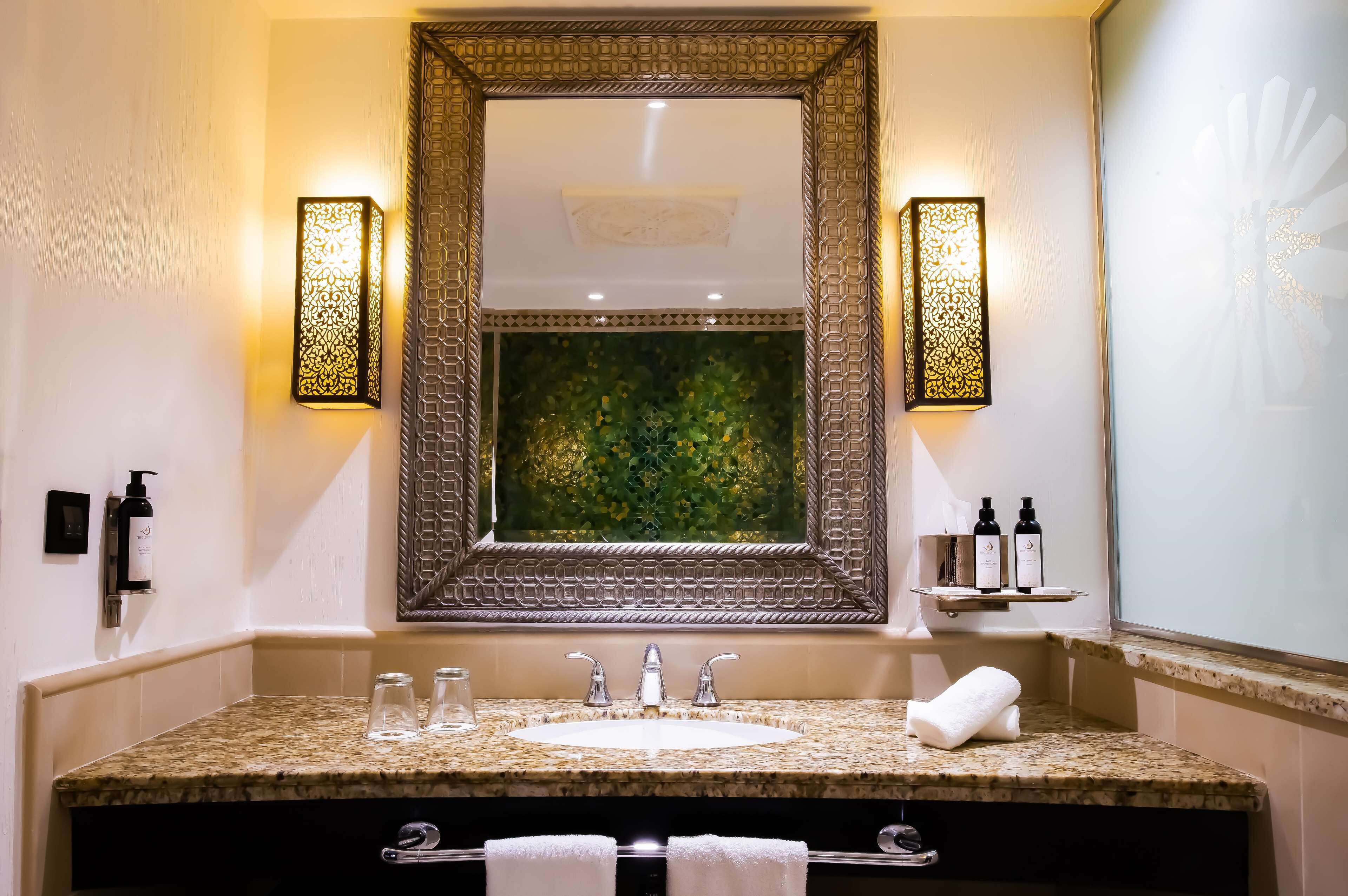 A bathroom with a large mirror, two wall-mounted lights, a sink with a granite countertop, and a decorative wall panel.