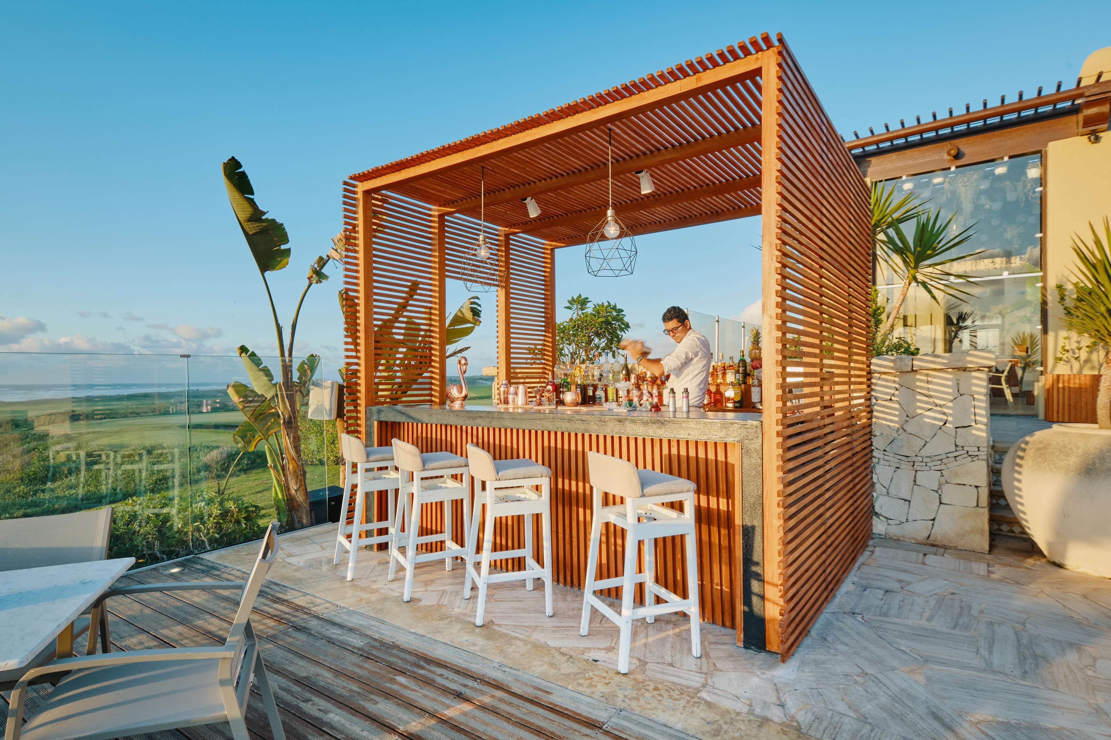 A wooden outdoor bar with white bar stools, a chef preparing food, and a scenic view.