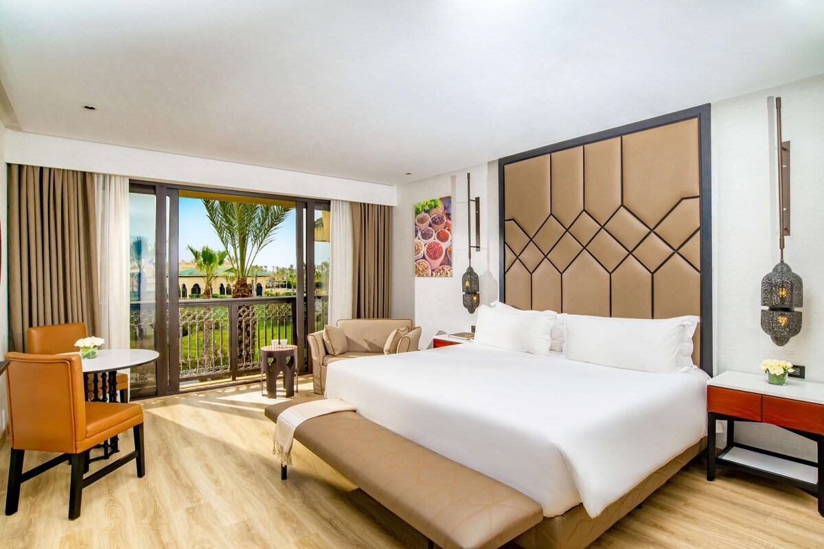 A hotel room with a large bed, a sofa, a small table, and a bench. There is a balcony with a view of greenery and a palm tree.