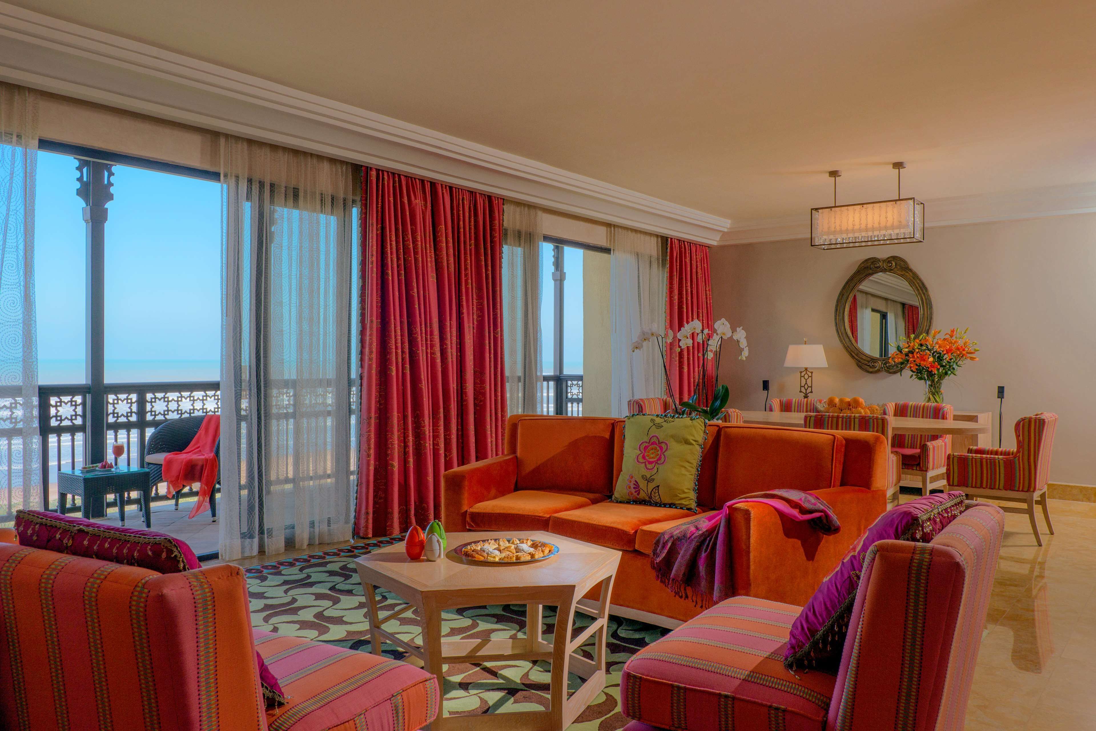 A spacious living area with a large sofa, armchairs, and a dining table. There is a balcony with a view of the sea.