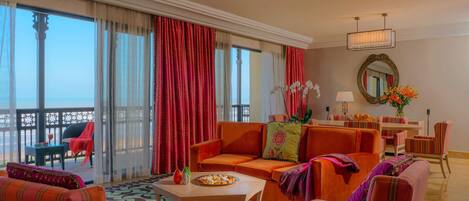 A spacious living area with a large sofa, armchairs, and a dining table. There is a balcony with a view of the sea.