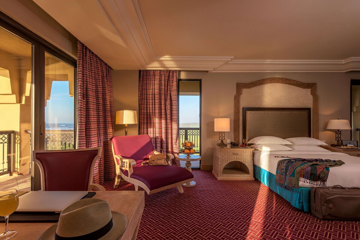 A hotel room with a bed, a chair, a table with a glass of wine, and a view of the ocean.