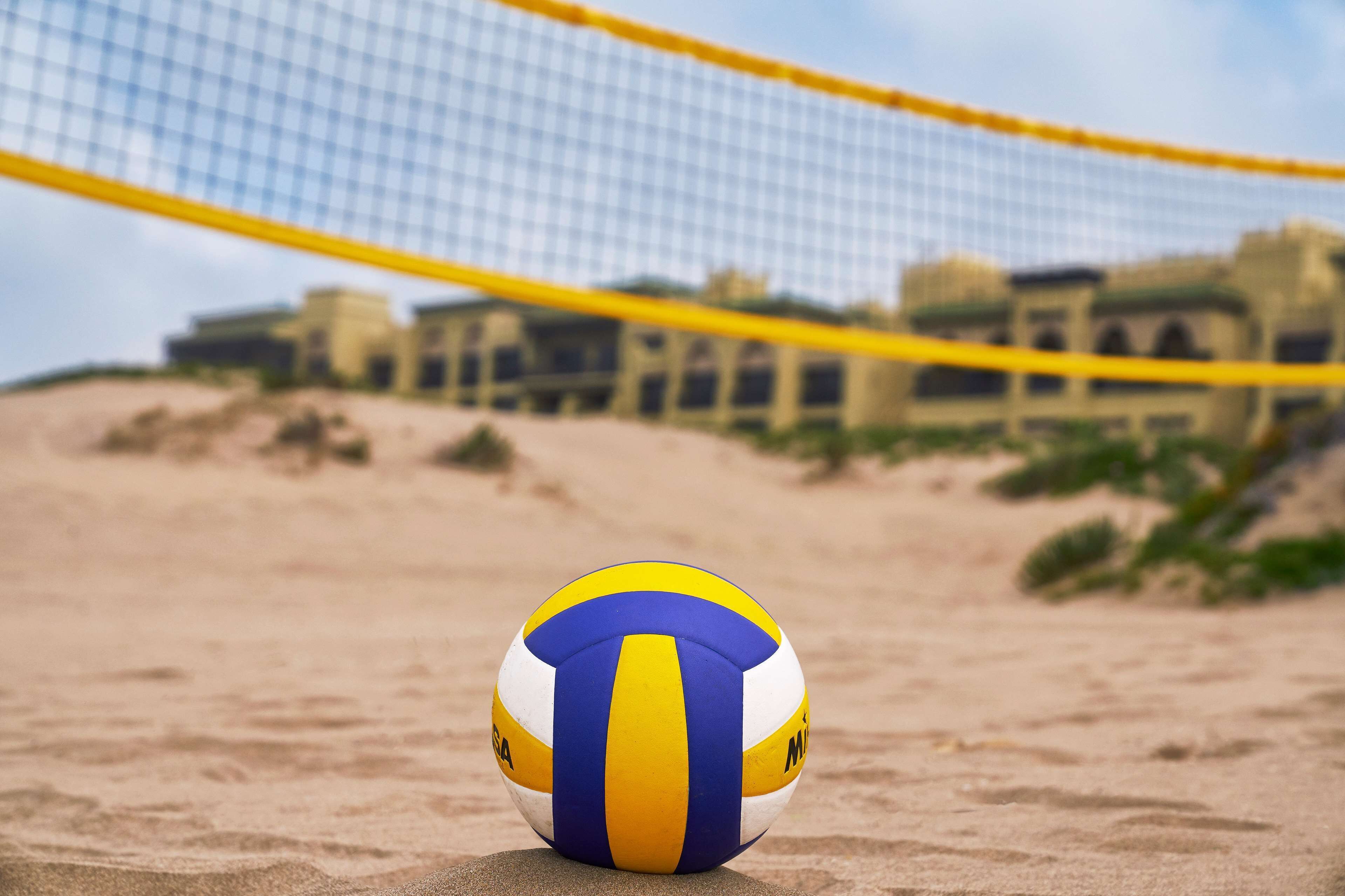 Ein Strandvolleyball auf sandigem Untergrund, im Hintergrund ein Netz und in der Ferne Gebäude.