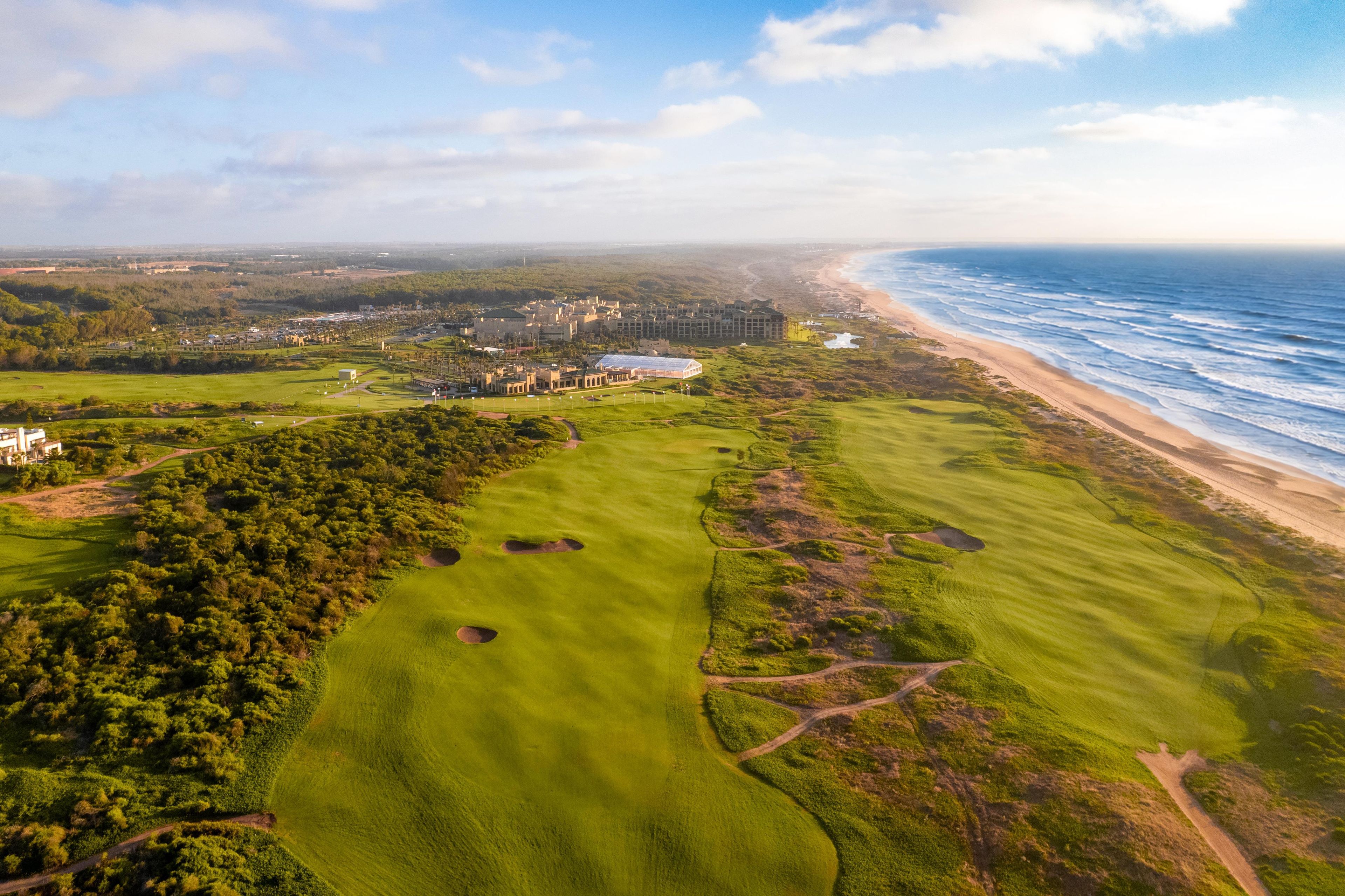 An aerial view of a coastal golf course with a beach and ocean in the background.