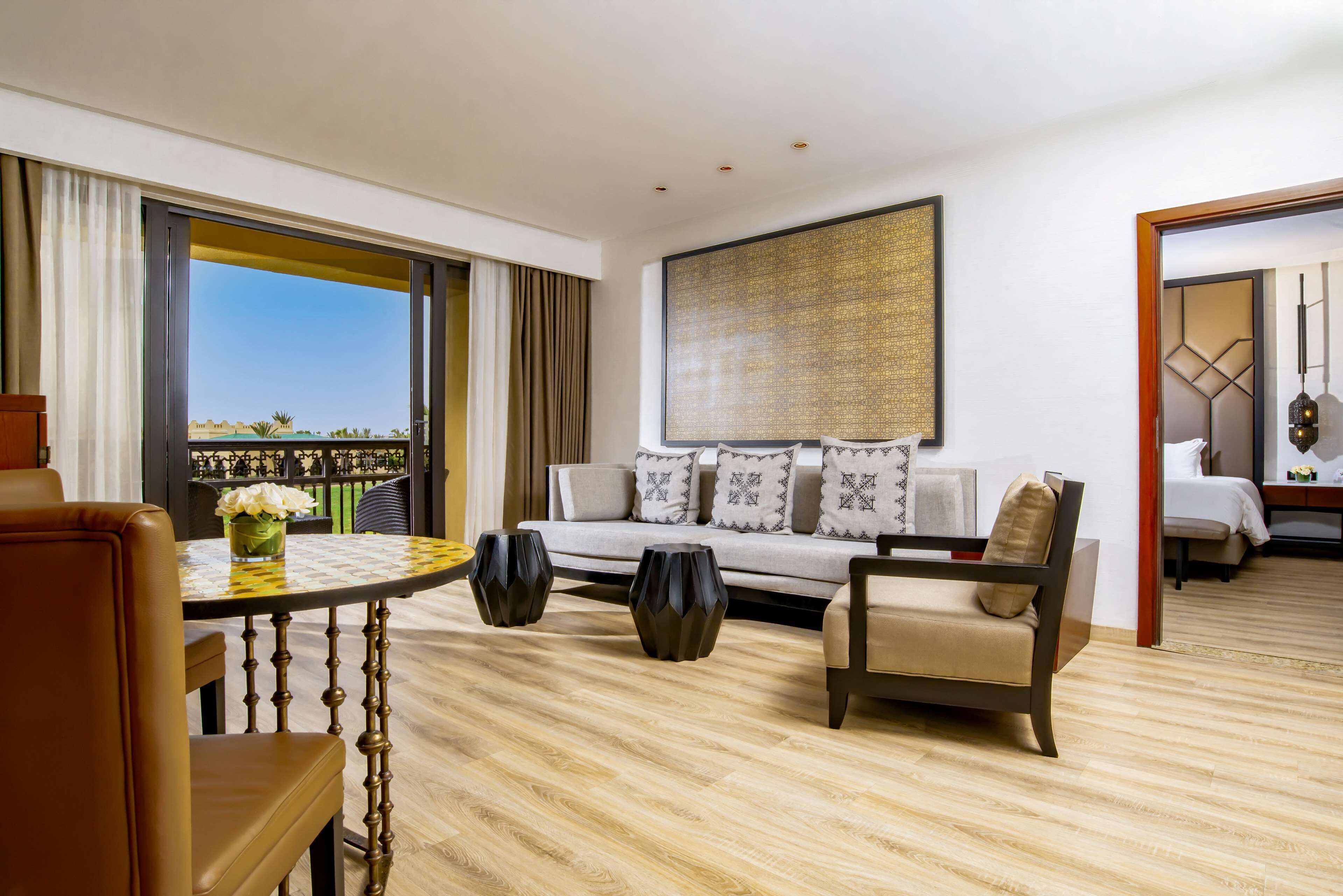 A hotel room with a sofa, armchairs, a dining table, and a large window offering a view.