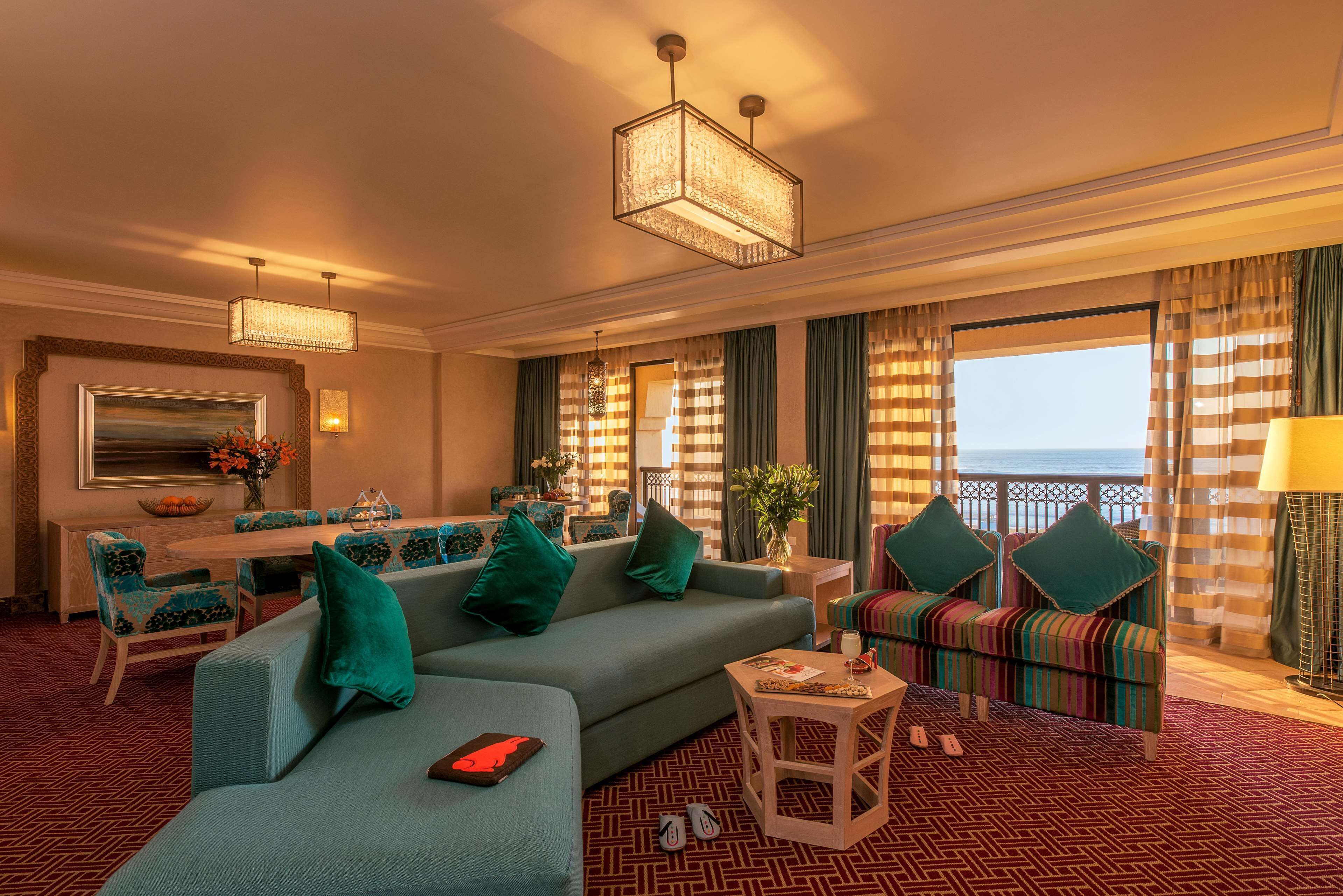 A hotel room with a sofa, chairs, a dining table, and a view of the sea.