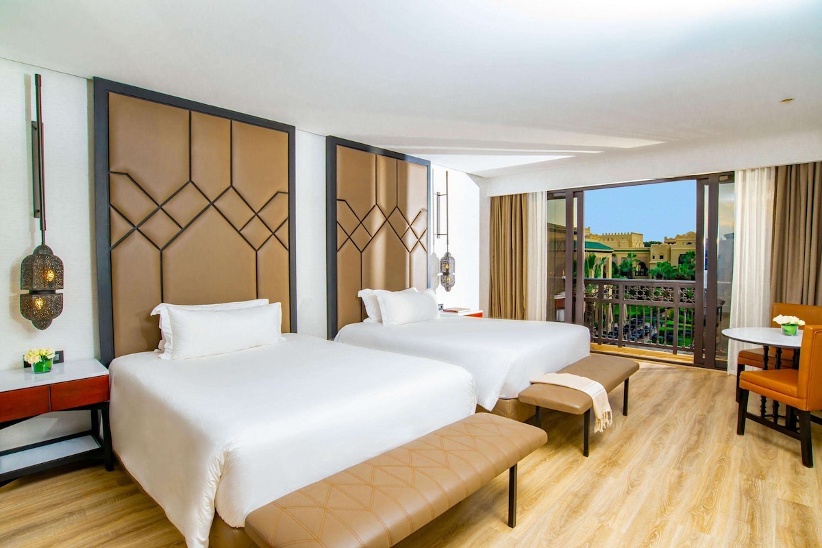 A hotel room with two beds, a desk, and a balcony with a view.