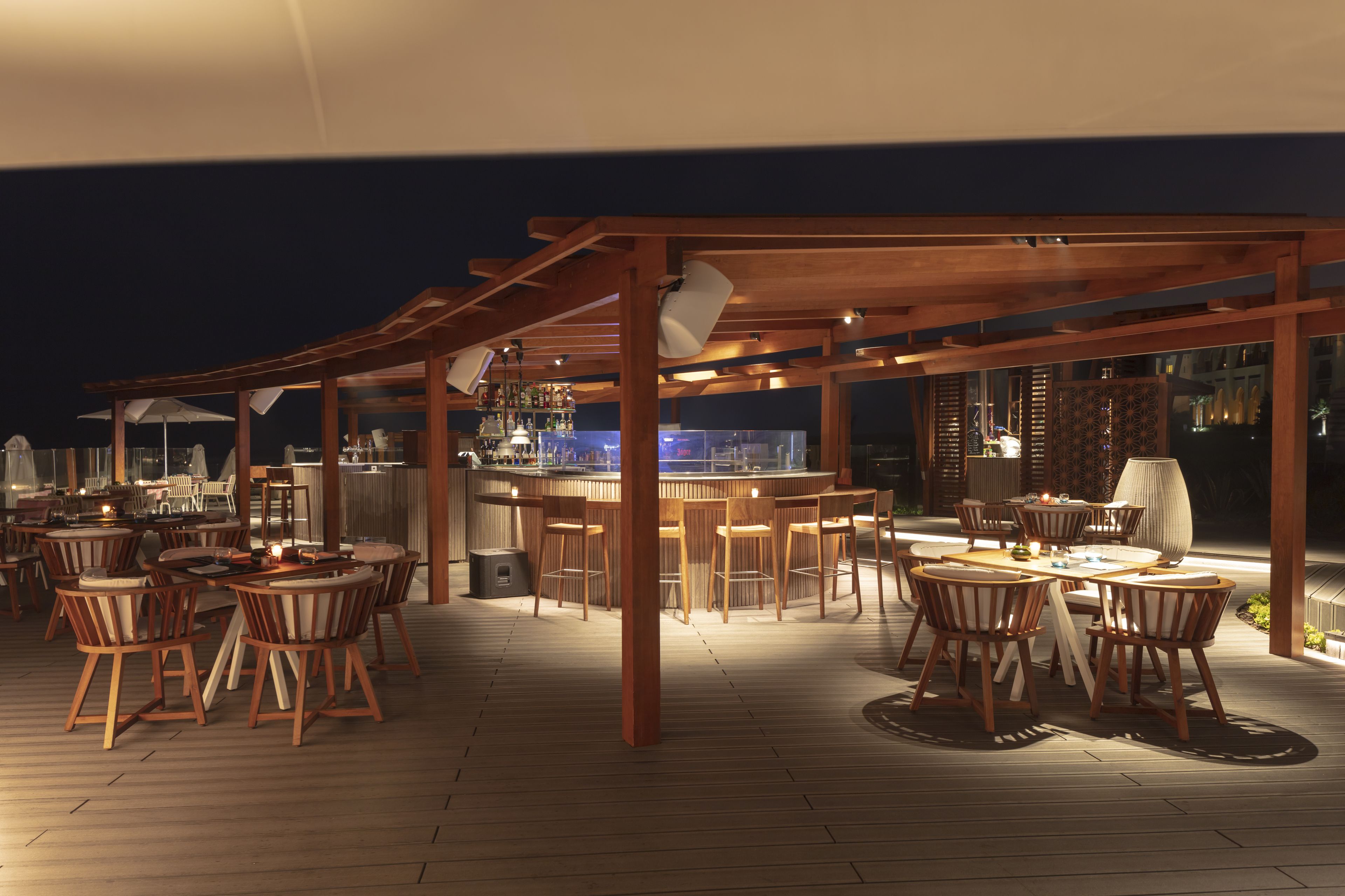 Un área de comedor al aire libre con mesas y sillas de madera, una barra y una estructura de pérgola.