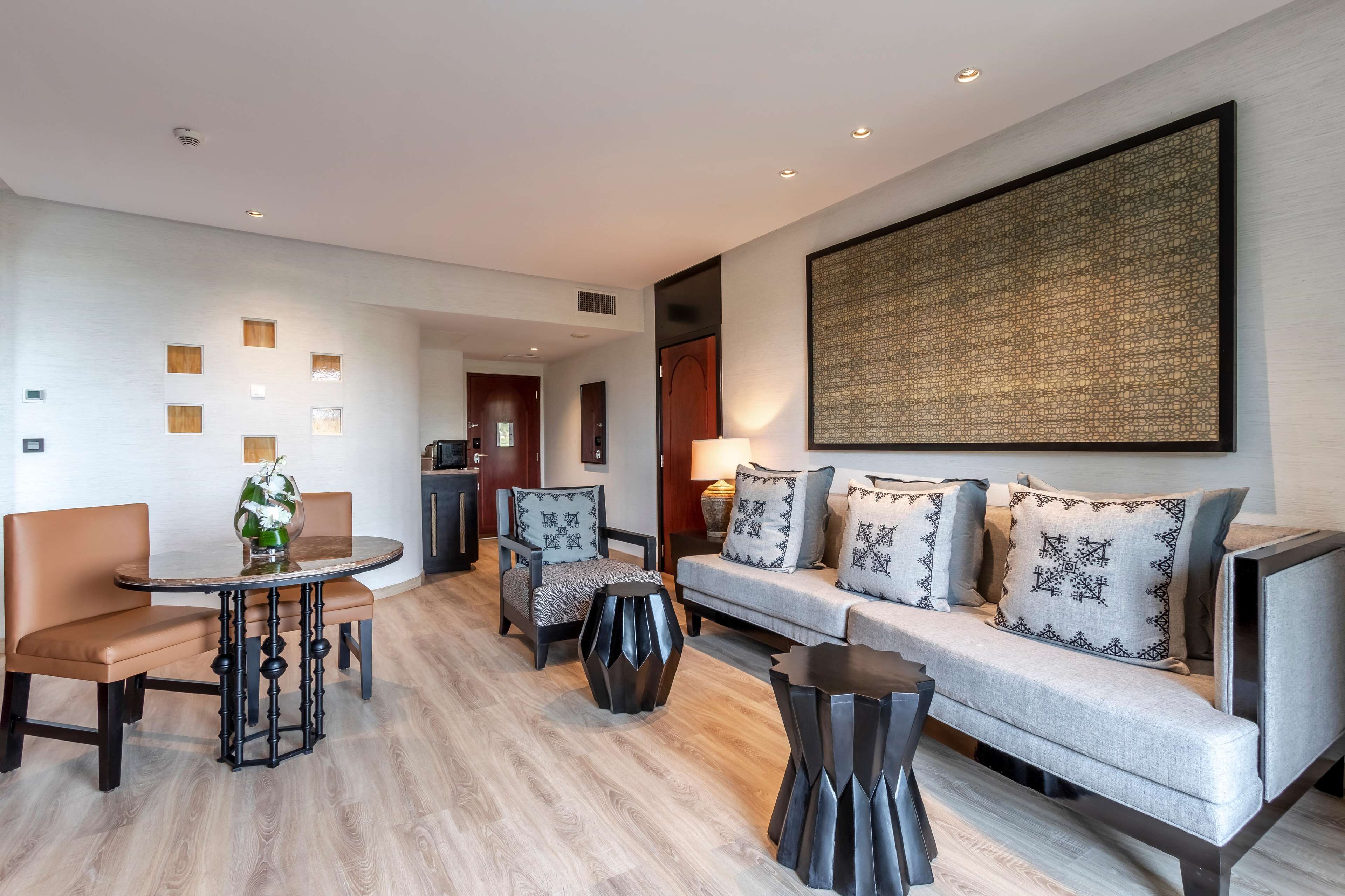 A modern hotel room with a sofa, chairs, a round table, and a large abstract artwork on the wall.