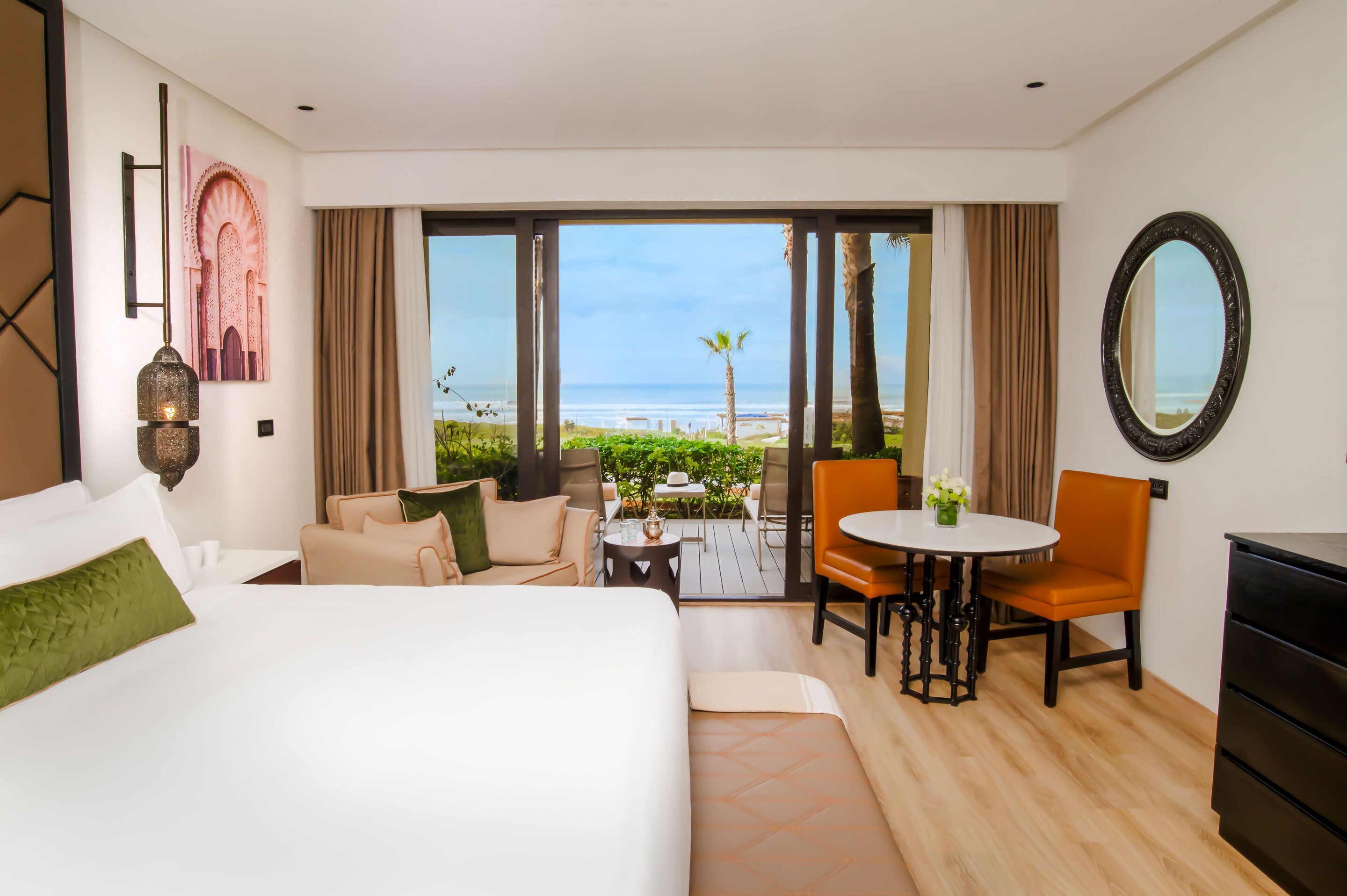 Habitación de hotel con una cama grande, zona de estar con una mesa y sillas, vista a la playa y un espejo grande en la pared.