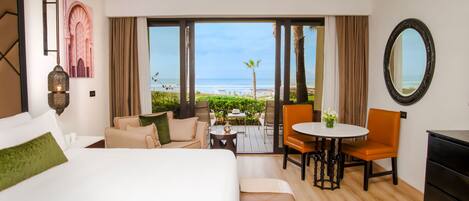 Habitación de hotel con una cama grande, zona de estar con una mesa y sillas, vista a la playa y un espejo grande en la pared.