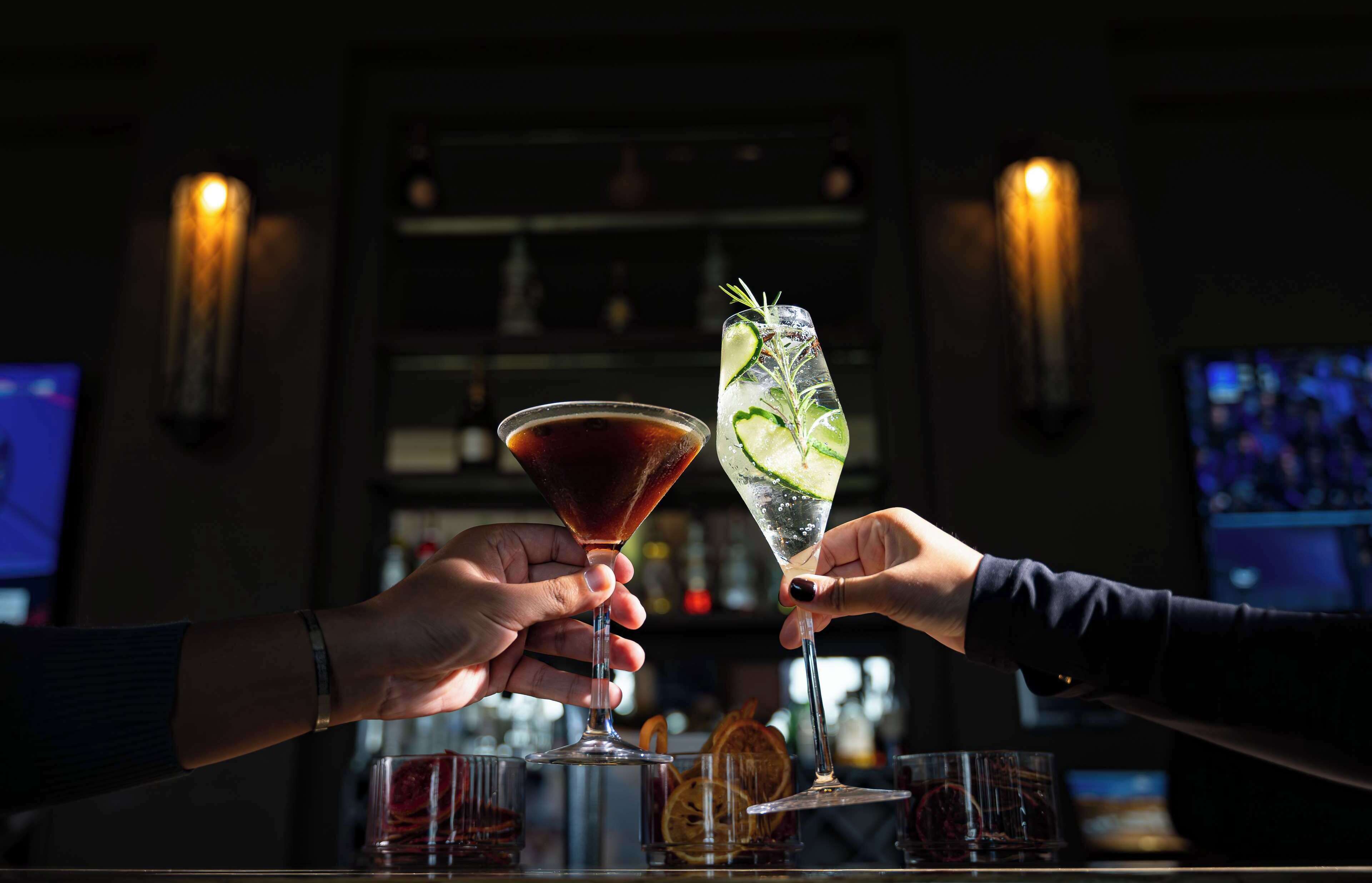 Two hands holding cocktail glasses at a bar with a dimly lit background and a television screen.