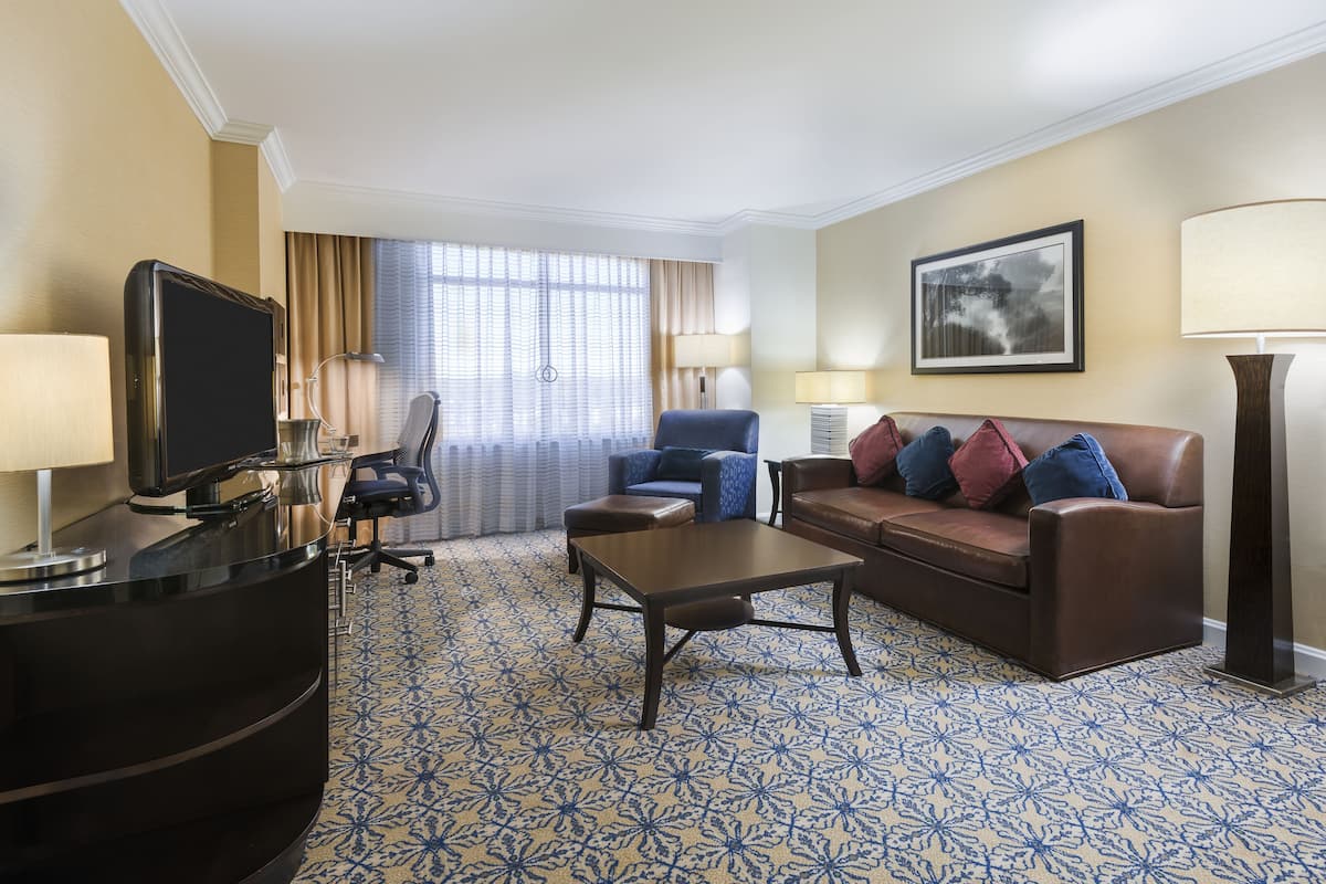 A hotel room with a desk, a brown leather sofa, a blue armchair, a coffee table, and a patterned carpet.