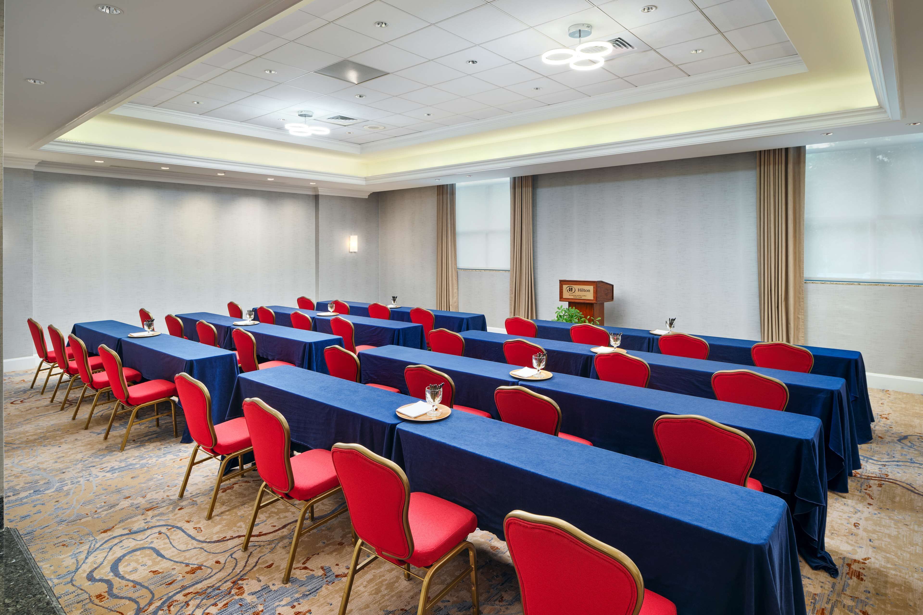 Un salón de conferencias con filas de mesas y sillas, un atril y una pantalla para proyector.
