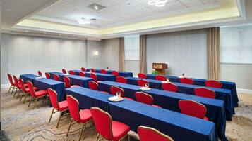 Un salón de conferencias con filas de mesas y sillas, un atril y una pantalla para proyector.