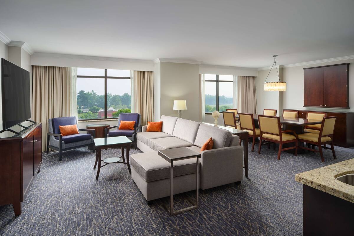 A hotel room with a sectional sofa, armchairs, a dining table with chairs, a TV, a lamp, and large windows with curtains.