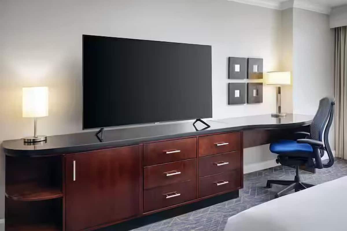 A hotel room with a flat-screen TV, a wooden desk with drawers, a chair, and a bed.
