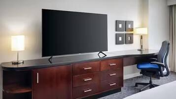 A hotel room with a flat-screen TV, a wooden desk with drawers, a chair, and a bed.