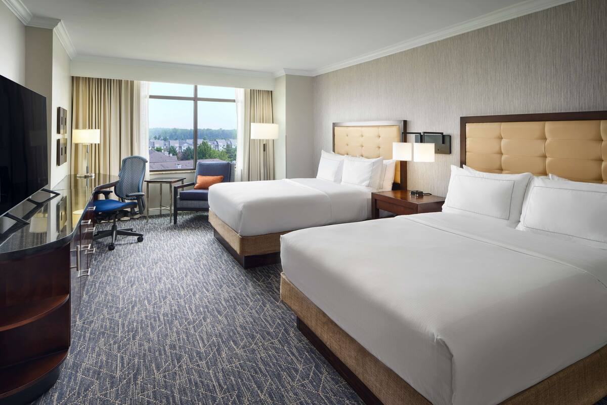 A hotel room with two beds, a desk, a chair, a TV, and a large window with curtains.