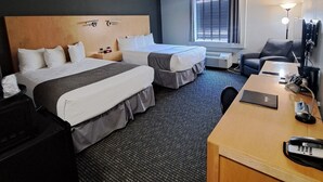 Quarto de hotel com duas camas, uma escrivaninha, uma cadeira, um abajur e uma janela com persianas.