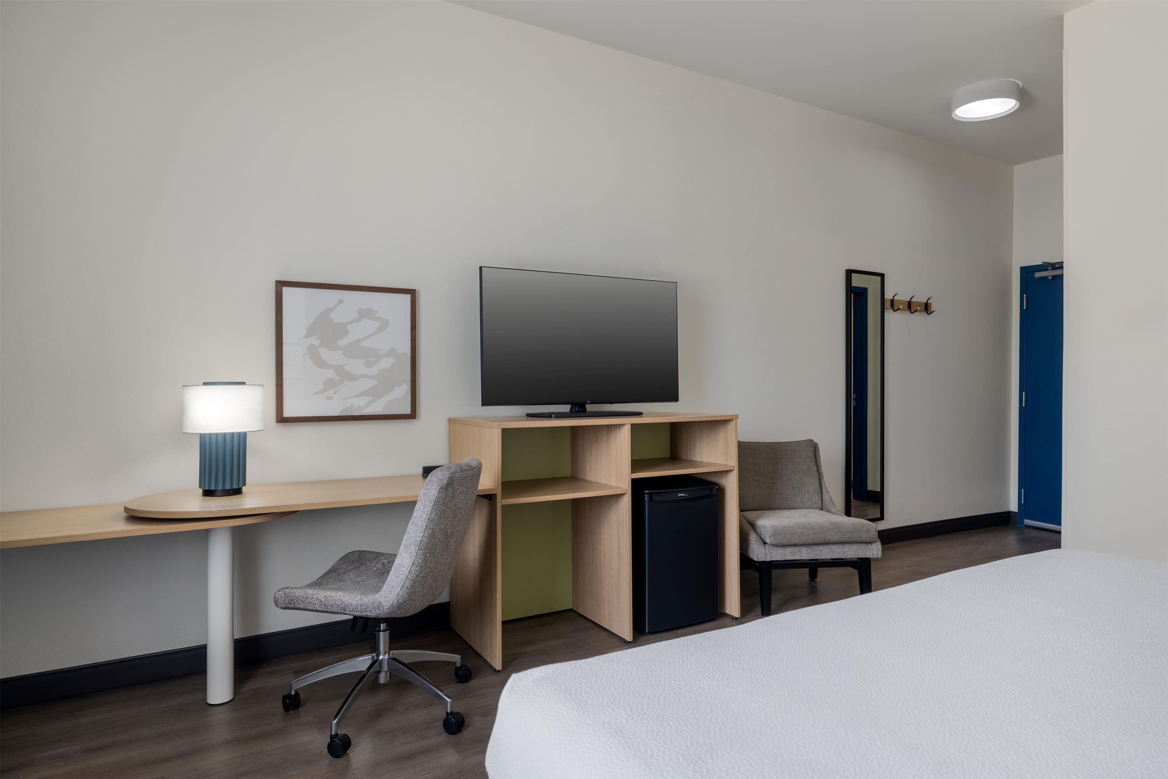 A hotel room with a bed, a desk with a computer, a chair, a television, and a small refrigerator.