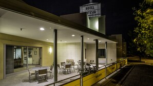 Exterior de um hotel com área de estar coberta, um letreiro indicando "HOTEL ARTESIA" e uma barreira amarela ao longo da calçada.