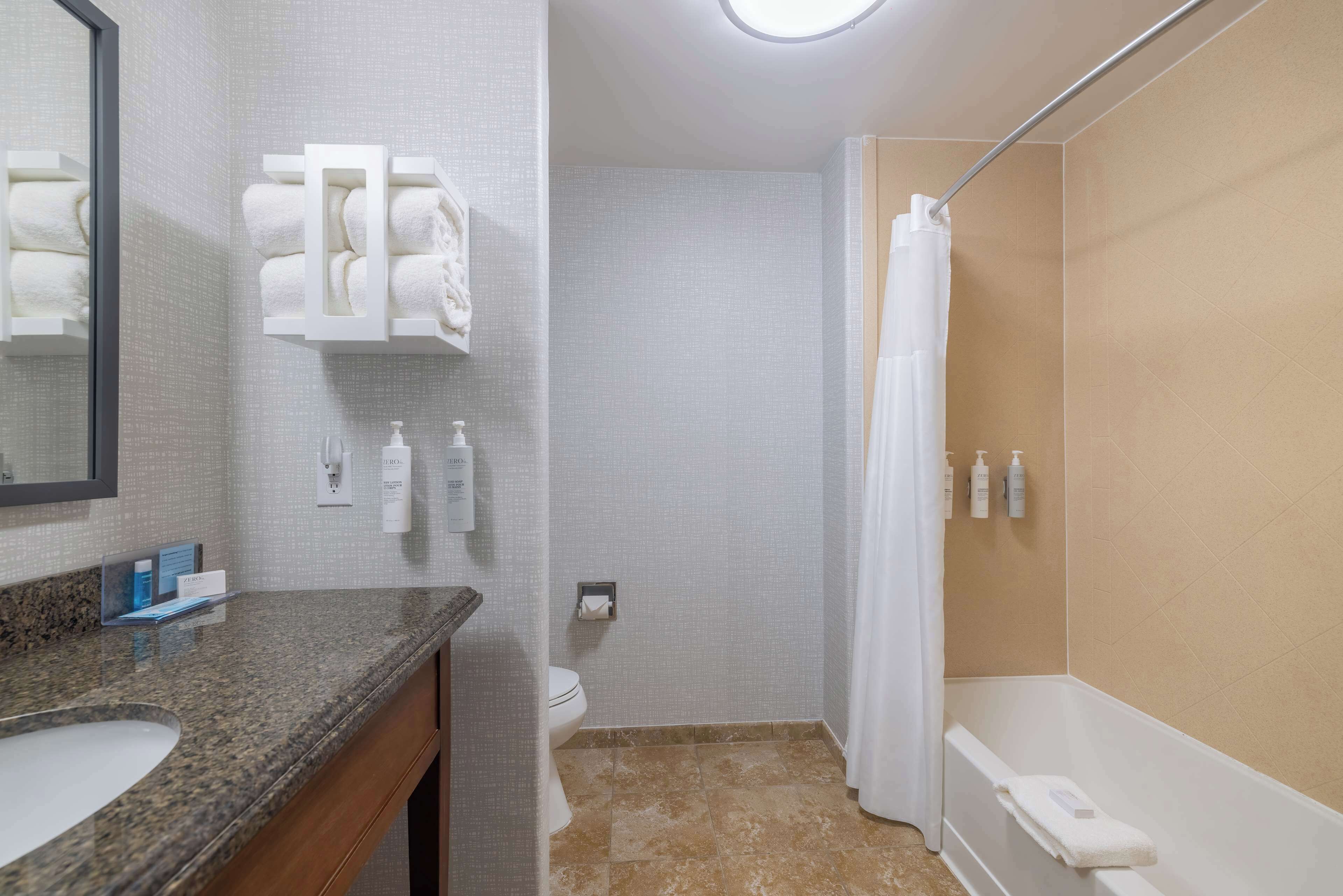A bathroom with a granite countertop sink, a toilet, a shower with a curtain, and a towel rack with folded towels.