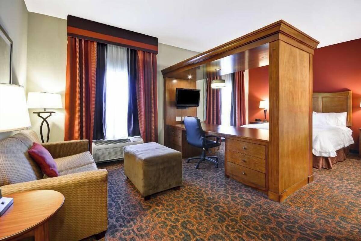 Une chambre d’hôtel comprenant un lit, un bureau, une chaise, un canapé, un pouf, une télévision et une lampe.