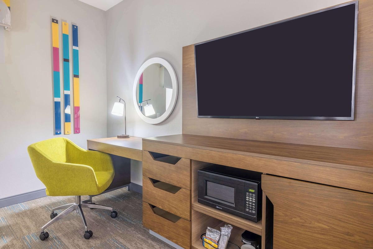 Une chambre d’hôtel avec un bureau en bois, une chaise jaune, un miroir rond, un téléviseur à écran plat et un four à micro-ondes.