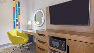 A hotel room with a wooden desk, a yellow chair, a round mirror, a flat-screen TV, and a microwave.