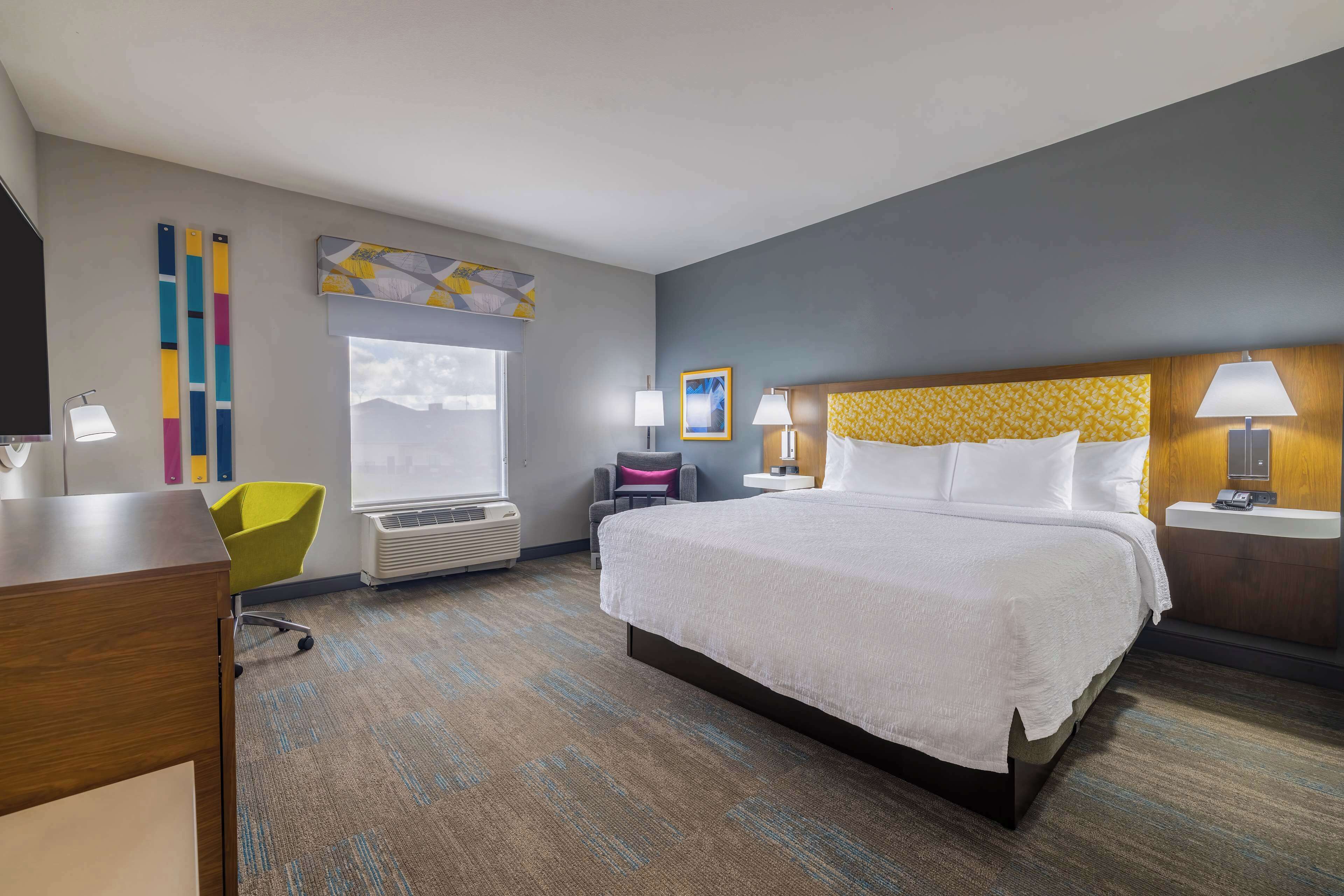 A hotel room with a large bed, a desk, a chair, a window, and a wall with artwork.