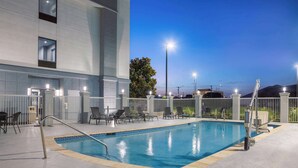 Una piscina interior con vistas a un edificio, un área vallada con sillas y un cielo despejado.