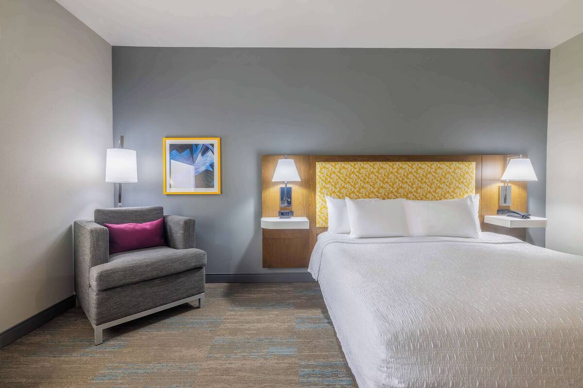 Une chambre d’hôtel avec un lit, un fauteuil gris agrémenté d’un coussin rose, deux tables de chevet avec des lampes, un tableau encadré au mur et une tête de lit à motifs.