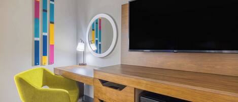 A hotel room with a wooden desk, a flat-screen TV, a microwave, a yellow chair, a round mirror, and colorful vertical blinds.