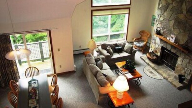 A cozy living room with a stone fireplace, a sofa, a dining table with chairs, and a large window providing a view of greenery outside. - Mount Snow Condominiums (West Dover)