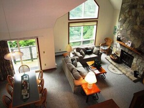A cozy living room with a stone fireplace, a sofa, a dining table with chairs, and a large window providing a view of greenery outside. - Mount Snow Condominiums (West Dover)