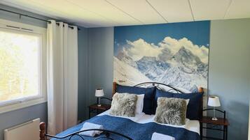 A bedroom with a mountain mural, a bed with blue bedding, two bedside tables with lamps, and a window with curtains.