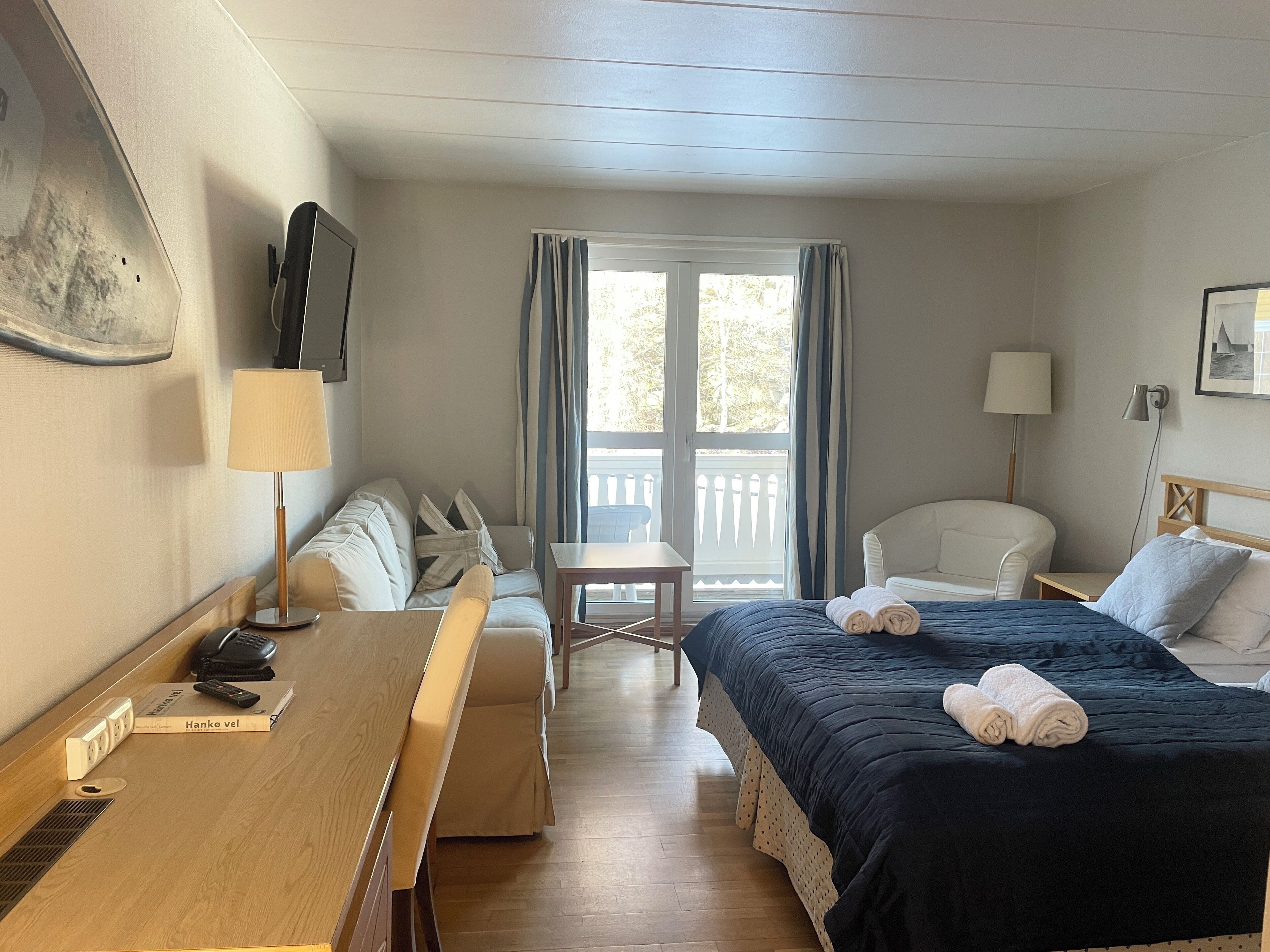 Une chambre d’hôtel avec un lit, un bureau, une chaise, une petite table, une lampe et une fenêtre avec des rideaux.