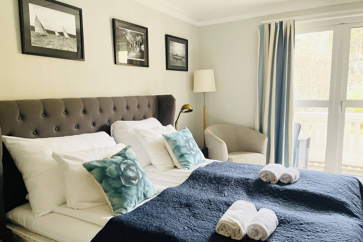 A neatly made bed with white and blue pillows, a dark blue quilt, and rolled towels. There are three framed pictures on the wall above the bed.