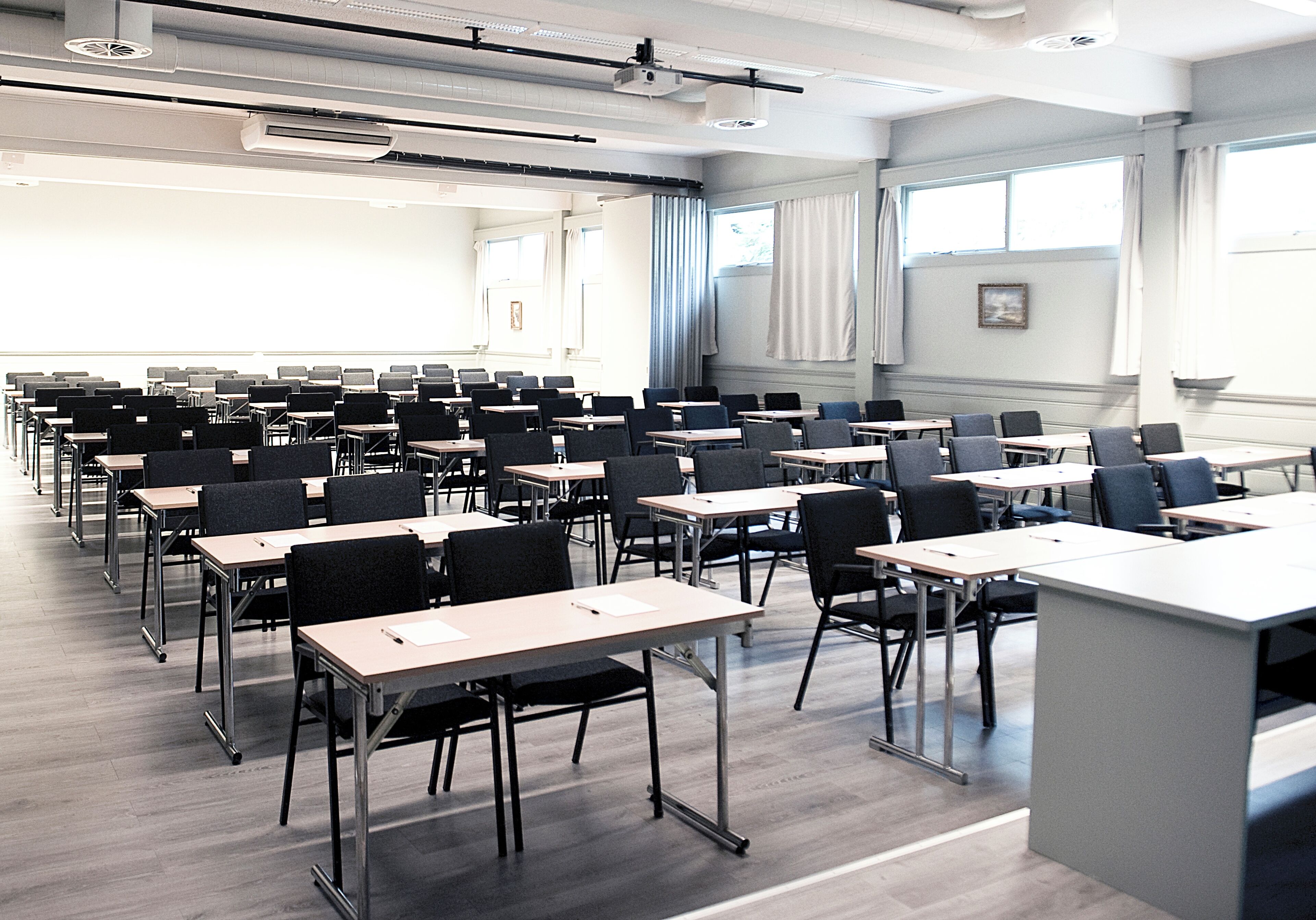 A classroom with rows of desks and chairs, large windows, and a projector on the ceiling.
