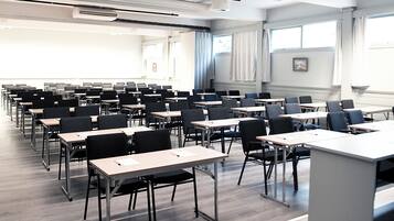A classroom with rows of desks and chairs, large windows, and a projector on the ceiling.