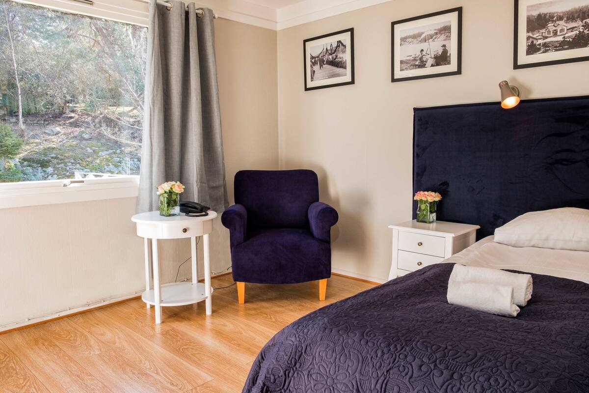 A bedroom with a dark headboard, a bed with a dark quilt, a small white side table with a vase of flowers, a purple armchair, and a window with a view of trees and a path.