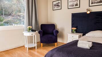 A bedroom with a dark headboard, a bed with a dark quilt, a small white side table with a vase of flowers, a purple armchair, and a window with a view of trees and a path.