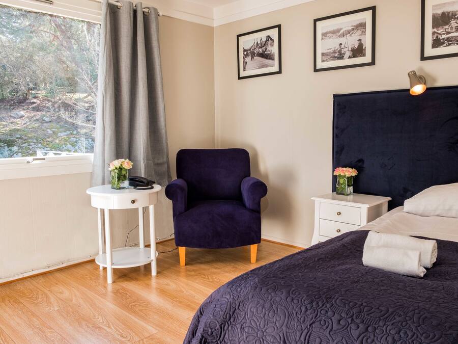 Un dormitorio con cabecera oscura, cama con colcha oscura, mesita de noche blanca con un jarrón de flores, sillón morado y ventana con vista a árboles y un sendero.