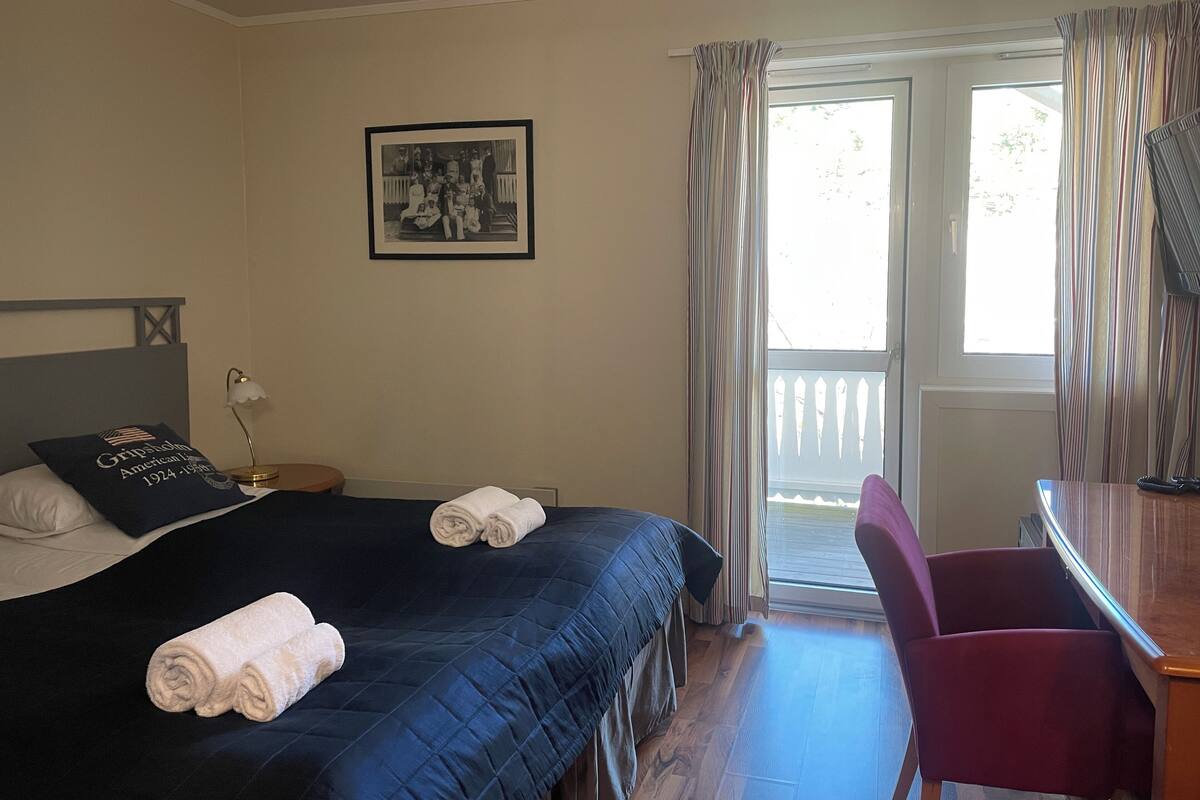 A hotel room with a bed, a desk, a chair, a framed photo on the wall, and a window with curtains.