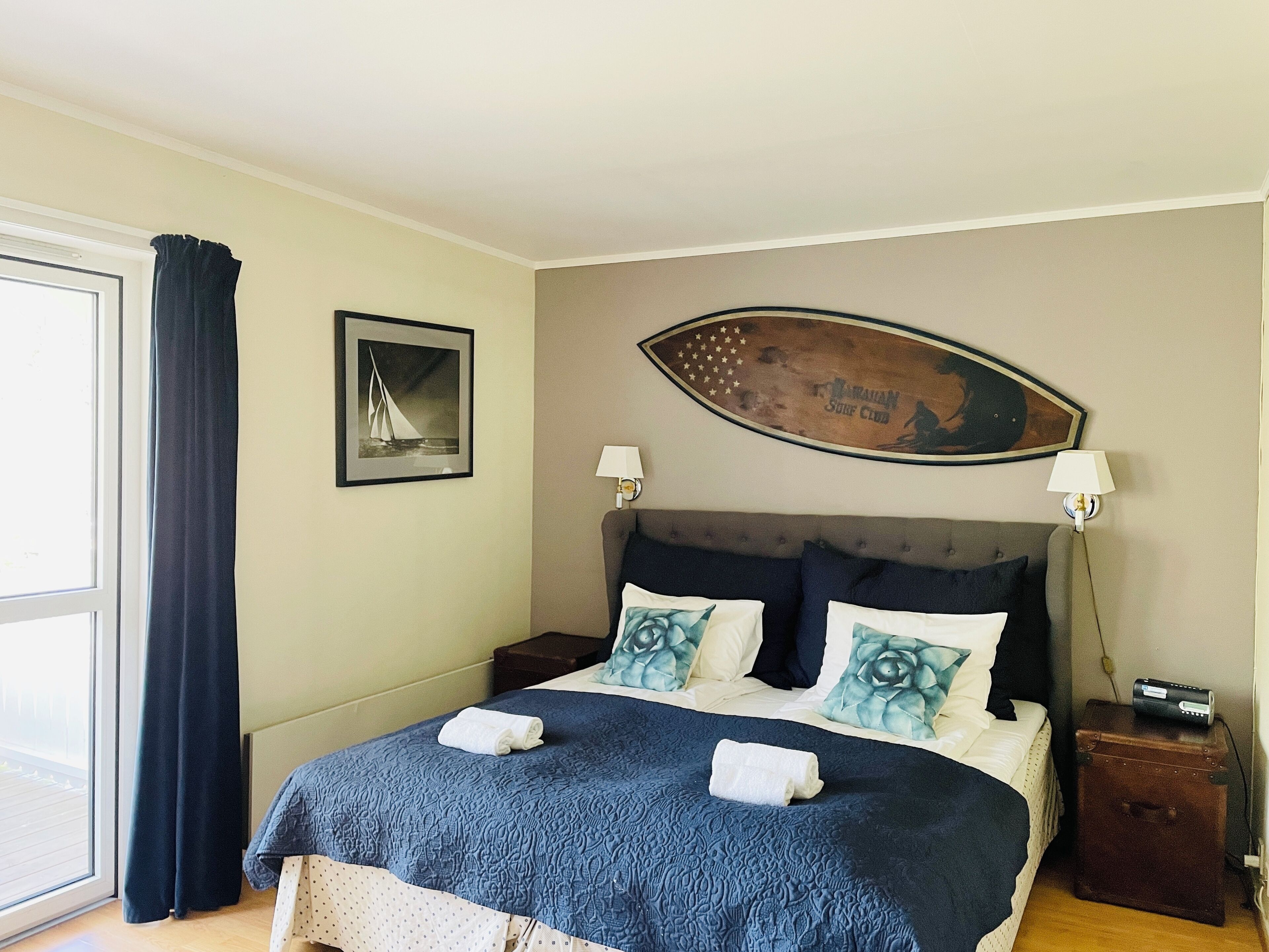 A neatly made bed with a blue quilt, two pillows, and a rolled towel. There is a surfboard-shaped picture above the bed, a framed picture on the wall, and a window with curtains.