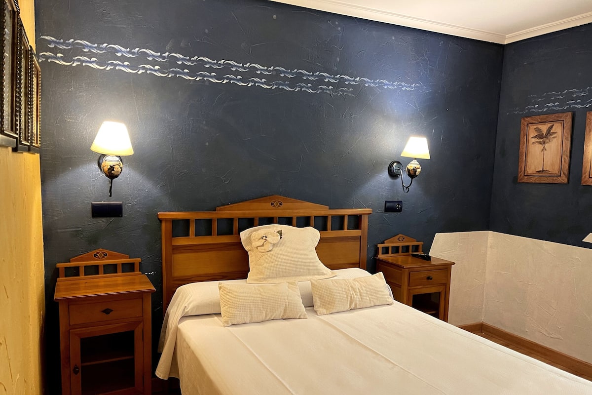 Una cama bien hecha con sábanas blancas y dos mesitas de noche, cada una con una lámpara. La habitación tiene paredes azul oscuro con detalles decorativos y dos cuadros enmarcados.