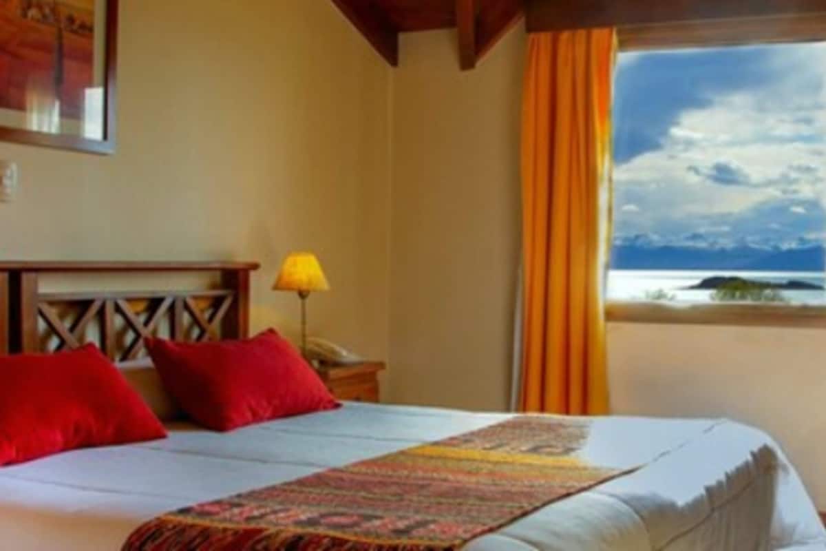A hotel room with a bed, red pillows, a patterned throw, a nightstand with a lamp, and a window with a view of the sea.